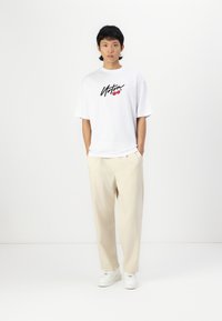 White oversized t-shirt with black and red graphic text, paired with beige wide-leg trousers and white sneakers. Plain background.