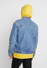 Denimjacka i ljusblått med en kontrasterande gul hoodie som syns under. Har traditionella sömmar och tryckknappar vid midjan.