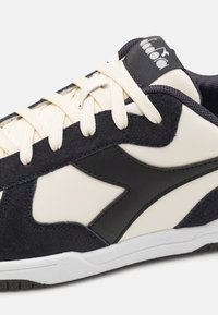 Sneaker con tomaia in pelle crema, accenti in suede blu scuro e logo nero. Presenta lacci piatti crema e una suola bianca testurizzata.