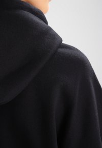Sudadera negra con capucha de textura suave, que presenta un corte holgado y una capucha prominente. Diseño minimalista sin logotipos visibles.