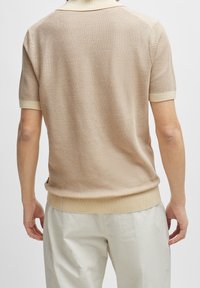 Beige kortärmad polo-tröja med en texturerad stickning, kontrasterande krage och ribbat fåll, bärs tillsammans med ljusa byxor.