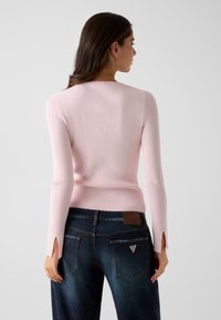 Růžový žebrovaný svetr s dlouhými rukávy a bočními rozparky, sladěný s tmavě modrými džínami s logovou nášivkou a klasickými zadními kapsami.