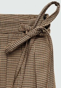 Tkanina s vzorom houndstooth v rjavaj in črni barvi z detajlom kravate na pasu. Material se zdi teksturiran in tkani, vsebuje vertikalne naborke.