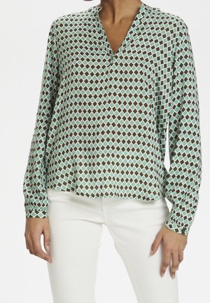 Personne portant un chemisier boutonné à manches longues avec un motif losange vert et marron, associé à un pantalon blanc sur fond blanc.