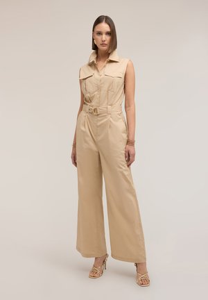 Mujer de pie, vestida con mono beige sin mangas con cinturón, pantalones de pierna ancha y tacones altos dorados con tiras sobre fondo neutro.