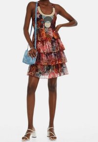 Robe à volants multicolores avec des motifs floraux, décolleté rond et détails en perles. Associée à un sac bandoulière bleu clair et des sandales blanches.