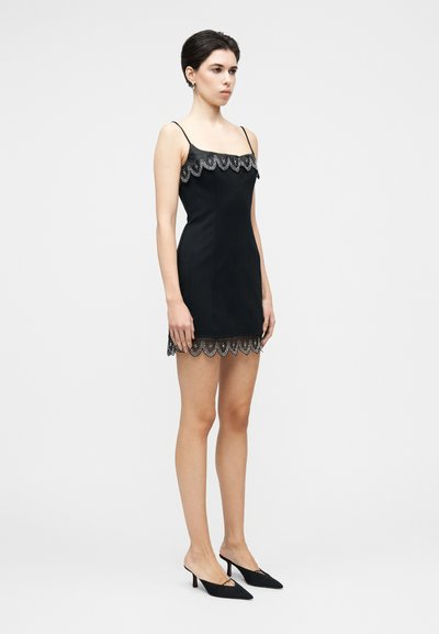 Melnā mini kleita ar plānām siksniņām, viļņotu mežģīņu apdari apakšmalā un kakla izgriezumā, kā arī piegulošu dizainu. Kombinēta ar melnām, ar smailu purngalu, čībām.