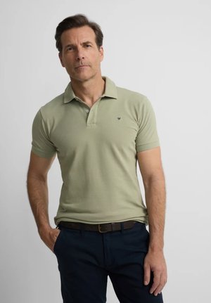 Hombre con cabello corto y castaño, vestido con camisa polo verde oliva y pantalones oscuros, de pie con una mano en el bolsillo contra un fondo liso.