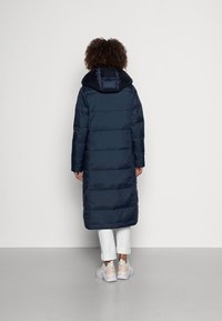 Dlouhý tmavě modrý péřový kabát s kapucí a prošívaným vzorem. Nošený s bílými kalhotami a světlými teniskami, pohled zezadu.