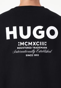 Black cotton t-shirt featuring large white text "HUGO" on the back. Additional white text includes "MCMXCIII", "REGISTERED TRADEMARK", and "SINCE 1993."