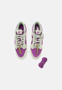 Purple suede sneakers with cream accents, featuring textured laces, a circular logo, and a spare matching shoelace positioned nearby.