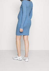 Femme en robe tricotée côtelée bleue et baskets blanches, debout de profil devant un fond blanc uni.