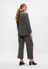 Sivi karirani sako s krojenim krojem i izrezom, spojeno s odgovarajućim hlačama širokog kroja, oboje izrađeno od teksturirane tkanine.