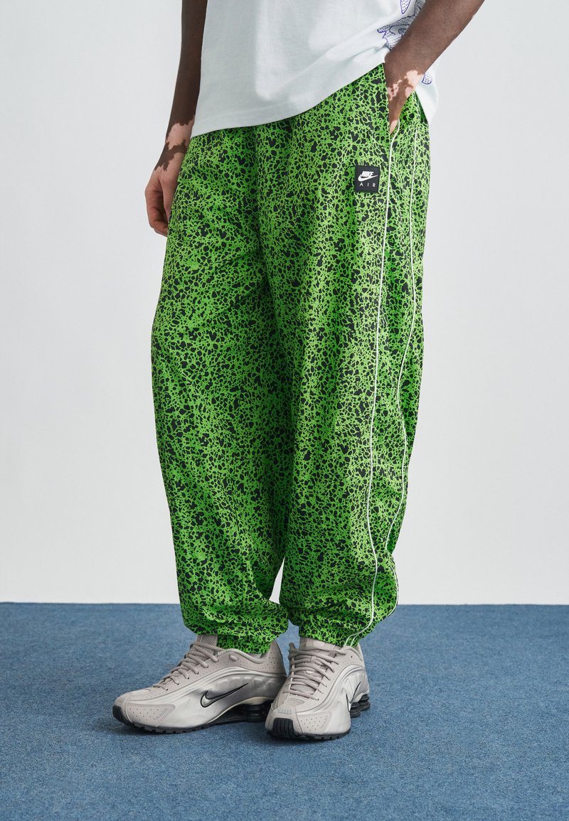 Zöld, mintás nadrág pöttyös dizájnnal, fekete logó folttal és fehér oldalsávokkal. Szürke tornacipővel párosítva.