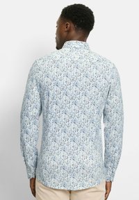 Mann von hinten, trägt ein hellblaues Hemd mit zartem Blumenmuster und beige Hosen vor einem einfarbigen Hintergrund.