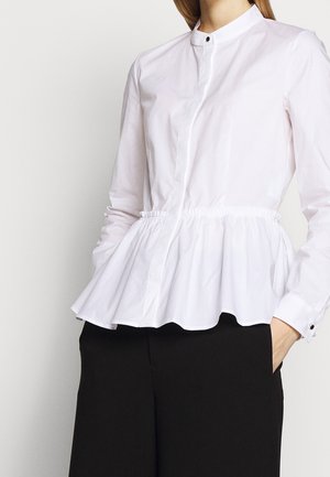 Woman wearing a white long-sleeve peplum blouse with buttoned cuffs and black pants, hand in pocket, against a plain background.