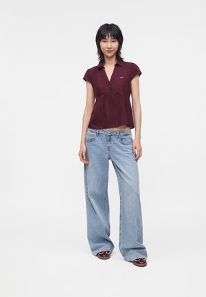 Junge Frau steht und trägt eine kurzärmelige weinrote Bluse, lockere hellblaue Jeans und weinrote Schuhe vor einem schlichten Hintergrund.