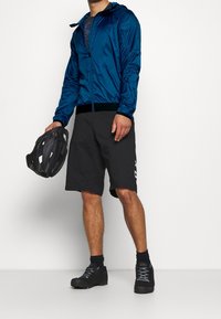 Man i blå jacka, svarta shorts och svarta skor som håller en svart cykelhjälm mot en vit bakgrund.