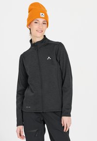 Grå zip-jacka med en strukturerad yta, med en vit logotyp på bröstet. Bärs med en orange stickad mössa. Svarta byxor med sidofickor.