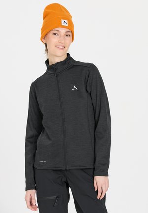 Grå zip-up jakke med en tekstureret overflade, der har et hvidt logo på brystet. Bæret med en orange strikket hue. Sorte bukser med sidelommer.