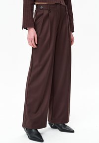 Persoon die een high-waist brede pantalon in bruin draagt, een cropped bruin pinstripe colbert en zwarte enkellaarsjes met puntige neus, staand tegen een witte achtergrond.