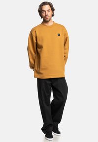 Sudadera de color mostaza con cuello redondo de canalé, corte oversize, bolsillos laterales y un pequeño parche con logo negro, combinada con pantalones anchos negros.