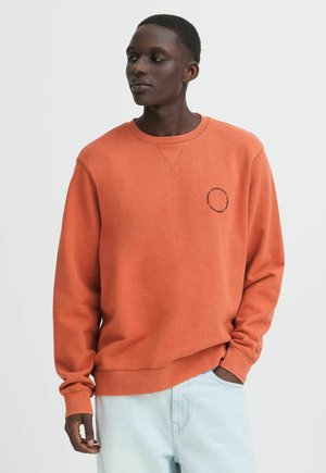 Jonge man draagt een oranje sweatshirt met cirkelvormige tekst op de borst en lichtblauwe spijkerbroek, staand met één hand in de zak.