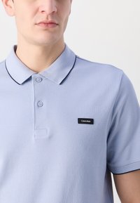 Világoskék poloing, textúrált anyagból készült, két gombbal és fekete gallérral. Ezen kívül található egy téglalap alakú fekete címke "Calvin Klein" logóval.