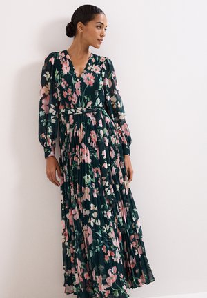 Frau trägt ein langärmeliges, dunkelgrünes, plissiertes Maxikleid mit rosa-weißem Blumenmuster und steht vor einer schlichten weißen Wand.