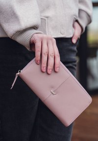 Pink leather wallet with a zip closure, featuring a gold accent logo, held in a hand, with a soft textured finish.