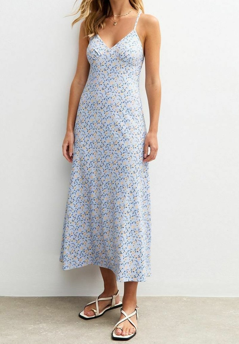 Femme portant une robe midi sans manches bleu clair avec un petit imprimé floral et des sandales plates beiges à brides, debout sur un sol gris.