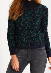 Maglione blu navy morbido con stampa leopardata turchese e nera, scollatura rotonda e polsini a costine. Abbinato a jeans grigi per un look casual.
