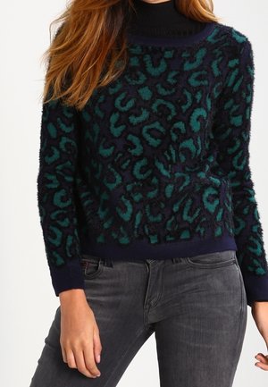 Luddig marinblå tröja med teal- och svart leopardmönster, rundad halslinning och ribbade ärmslut. Matchad med grå jeans för en avslappnad stil.