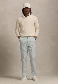 Pulover crem cu model tricotat în cablu, pantaloni bleumarin deschis, pantofi albi și o șapcă de baseball albă. Modelul stă cu mâinile în buzunare, pe un fundal neutru.