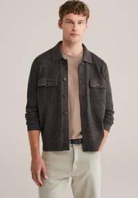 Grå ullkofta med krage, två bröstfickor och knappar. Buren över en ljusbrun skjorta och kombinerad med ljusa byxor.