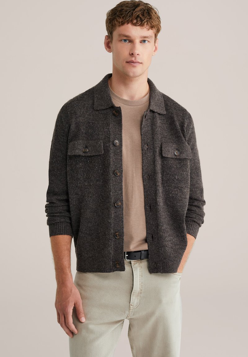 Grå ullkofta med krage, två bröstfickor och knappar. Buren över en ljusbrun skjorta och kombinerad med ljusa byxor.