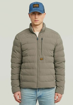 Homme portant une veste matelassée vert olive avec une poche poitrine zippée et une casquette en denim bleu, se tenant devant un fond léger uni.