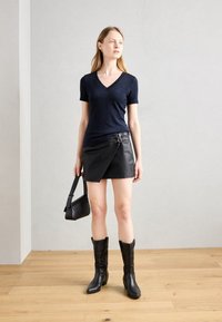 T-shirt marine à col en V, jupe ajustée en cuir noir avec accents de bouclerie, bottes noires jusqu'aux genoux et petit sac à épaule noir.