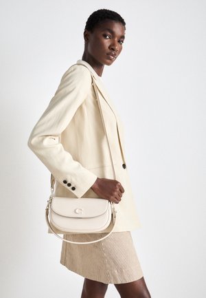 Femme portant un blazer crème et une jupe assortie avec un sac bandoulière crème sur un fond blanc uni.