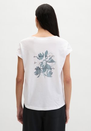 Femme aux longs cheveux bruns portant un T-shirt blanc avec un motif de feuilles bleu-gris et un texte en cursive dans le dos, assorti à un pantalon noir.