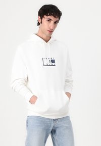 Sweat-shirt à capuche blanc avec une poche frontale et des poignets côtelés, arborant un logo "TOMMY JEANS" bleu et noir sur la poitrine. Design décontracté.