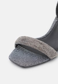 Sandale à talon tissée argentée scintillante avec une large bride pailletée et un design d'orteil carré texturé. Semelle intérieure noire mate visible.