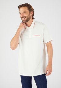 Camisa polo blanca con rayas rojas de acento en el collar y el bolsillo; de mangas cortas; hecha de una tela suave y transpirable. Llevada con jeans oscuros.