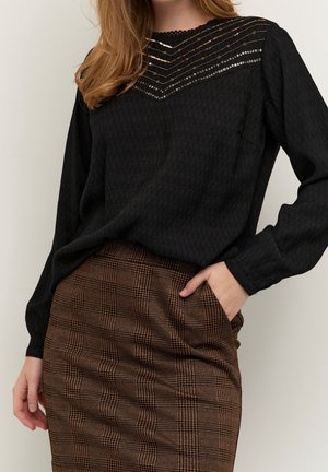 Kvinde iført sort langærmet top med metalliske striber, kombineret med høj talje brun ternet nederdel, hånd i nederdelslomme.