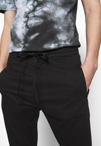 Pantalon noir à taille élastique avec cordon de serrage. Deux poches latérales, tissu doux et coupe ajustée. Porté avec une chemise grise et noire tie-dye.