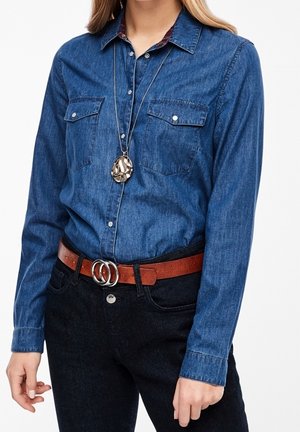 Denim-Hemd mit Knöpfen, zwei Brusttaschen, langen Ärmeln und einem karierten Kragen, getragen mit einem braunen Gürtel mit einer silbernen Doppelschnalle.