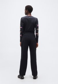 Άτομο που φοράει μαύρο μακρυμάνικο πουκάμισο με μοβ και μπεζ αφηρημένα σχέδια στα μανίκια και μαύρο παντελόνι, στέκεται γυρισμένο πλάτη σε λευκό φόντο.