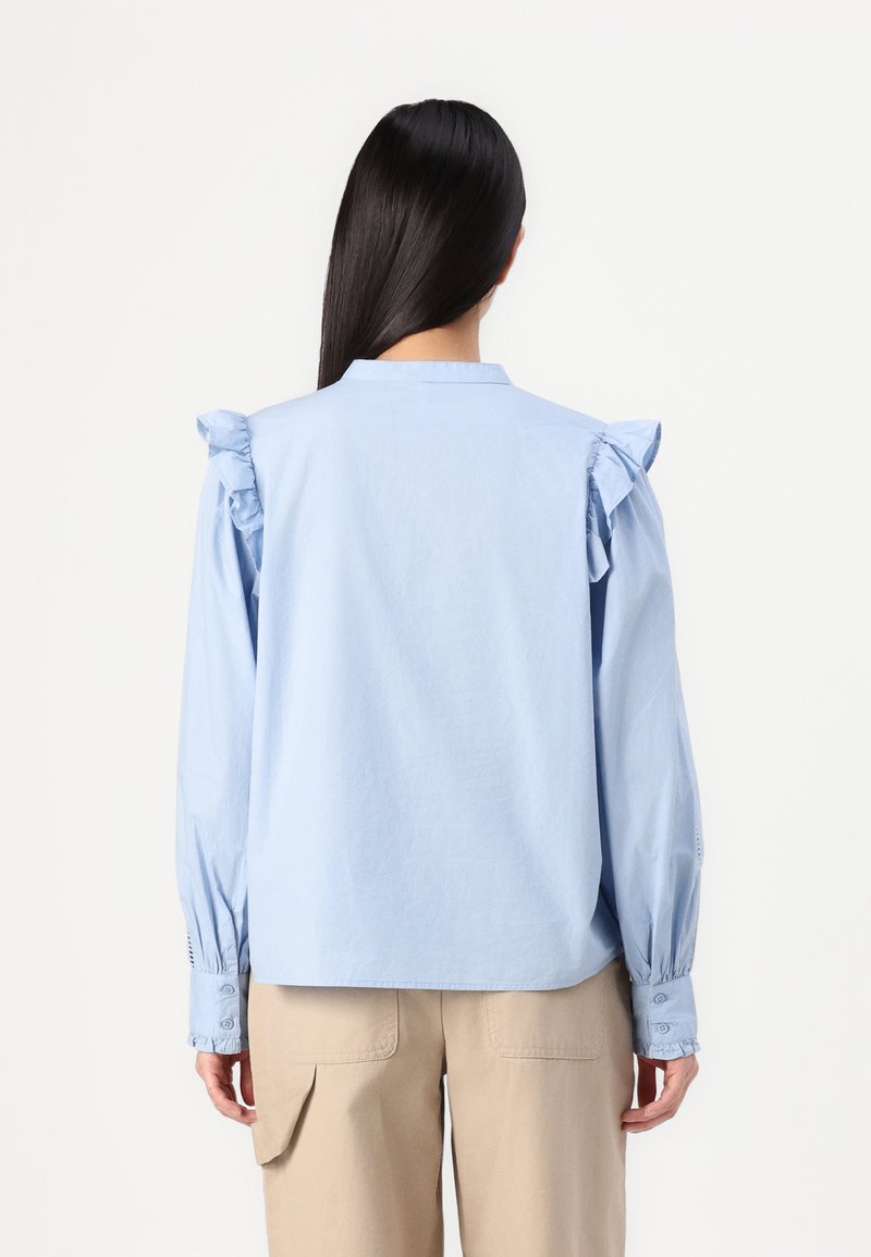 Chemise bleu clair à manches longues avec des accents de volants aux épaules, des poignets boutonnés et un col simple, associée à un pantalon cargo beige.
