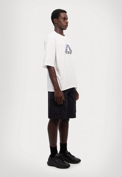 T-shirt en coton blanc avec un design graphique, associé à un short cargo bleu marine et des baskets noires. La tenue présente une coupe décontractée.
