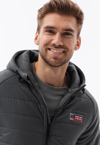 Chaqueta aislante sintética en gris oscuro con cierre frontal de cremallera, capucha forrada de polar y un detalle de parche en el pecho. Textura suave y ligera.